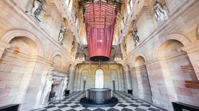 A Hall with a black and white checked floor and stone walls. Suspended from the ceiling is a large installation with long red fabric strips that connect at a central point.